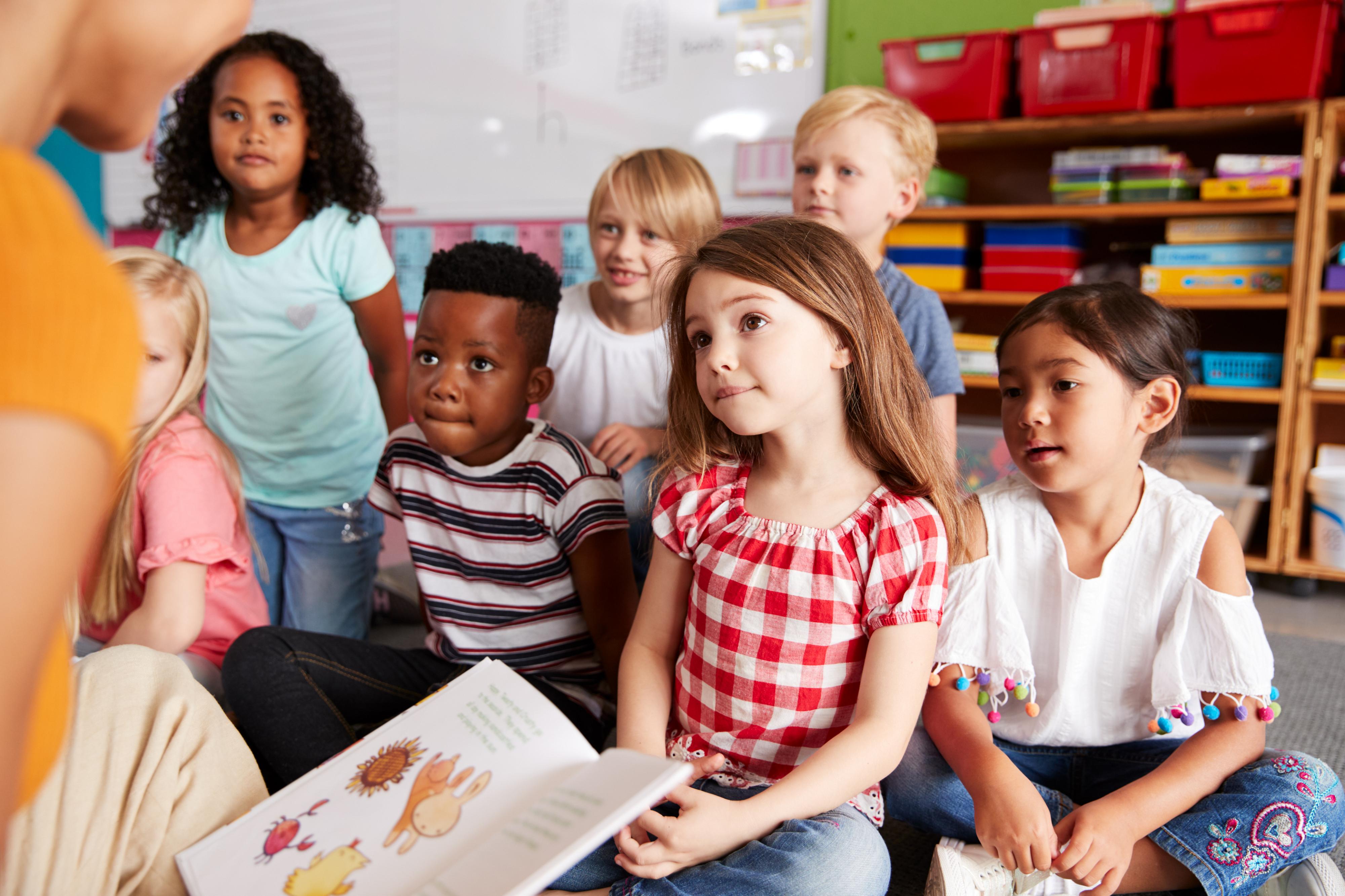 Group-Of-Elementary-School-Pupils-Sitting-On-Floor-Listening-To-Female-Teacher-Read-Story